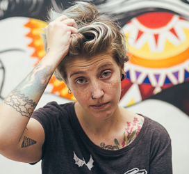 Person with tattoos and short hair standing in front of graffiti art with a tired expression