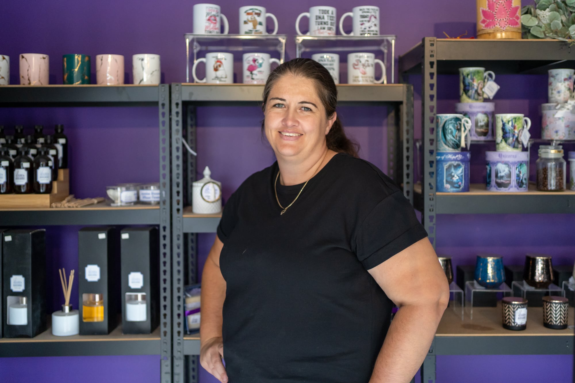 A women with her hair tied in a pony tail wearing a black T shirt standing in front of a purple wall with candles on shelves