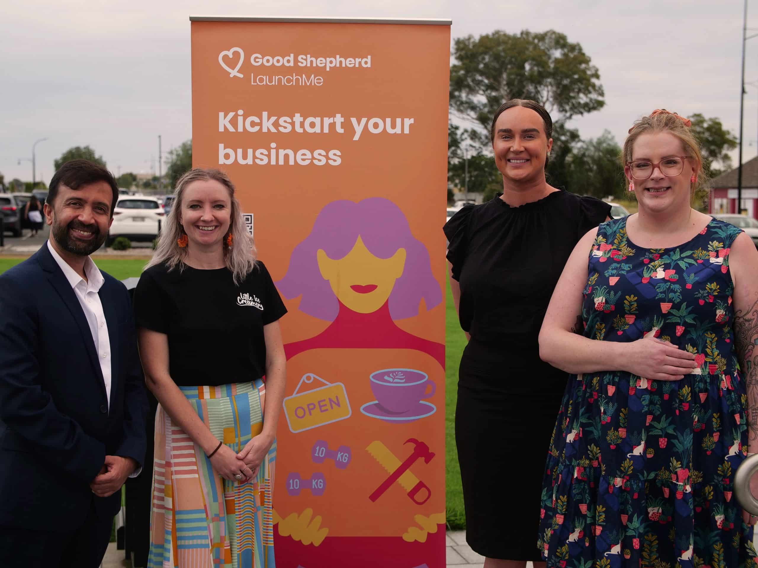 Left to right; a man in a suit standing next to three women in front of an orange sign which says Kickstart your business